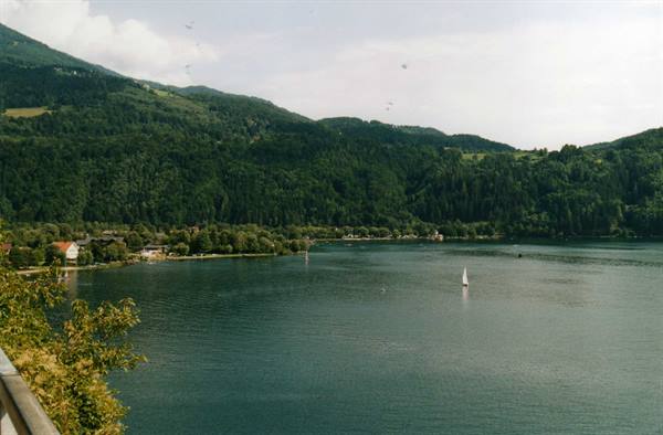 White sailboat on green lake surrounded by forested mountains and village