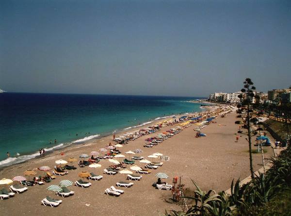 Beach with umbrellas and sunbeds, swimmers in the sea, buildings along the coast
