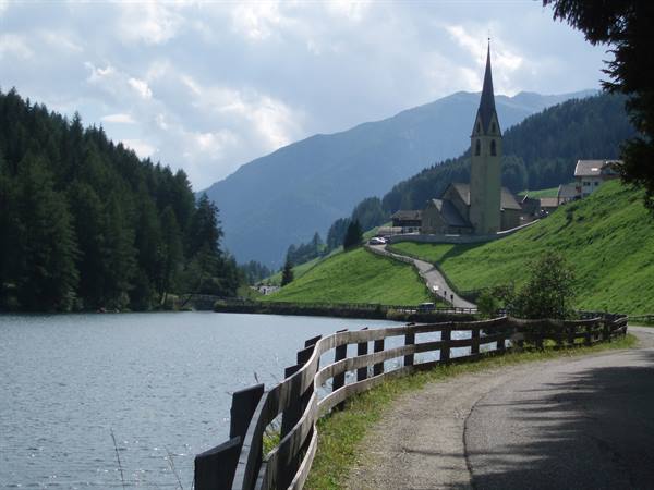 Dirt road with wooden fence beside alpine lake, church with tall steeple