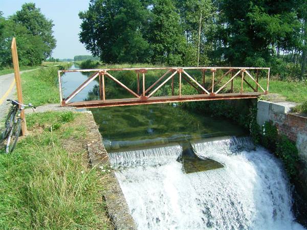 Red metal footbridge over a canal weir, bicycle parked on the grass