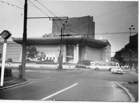 Modernist concrete theater at a city crossing, vintage bus and cars, black and white