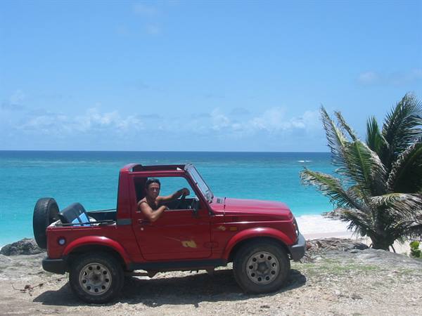 Man driving a red open-top jeep on sand by turquoise sea