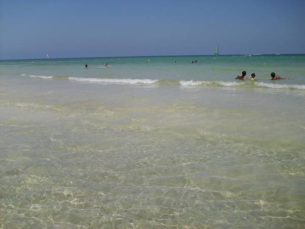 Swimmers in clear waves, green sail on horizon, sandy beach