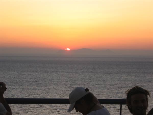 Sun sinking into the sea behind an island, people leaning on the railing