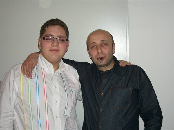 Two men embracing in front of a white wall, one with a cigar