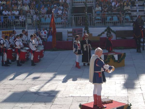 Drummers in medieval costume on a podium, crowd seated in town square stands