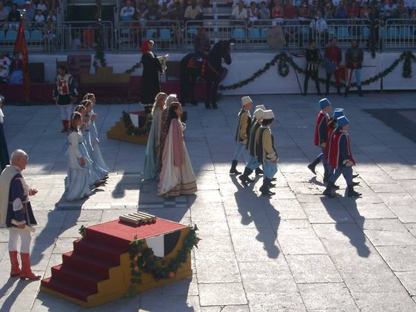 Medieval-costume parade in a stone square, children marching past a decorated platform