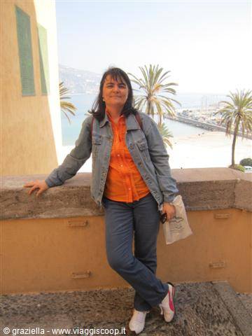 Woman in jeans and orange shirt leaning on a stone wall, palms and beach behind