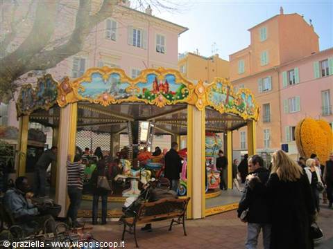 Ornate golden carousel with painted horses, tourists and benches in a square of pastel buildings