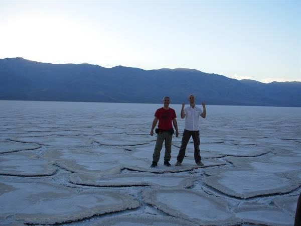 Two tourists pose on cracked salt at Badwater Basin, blue mountains behind
