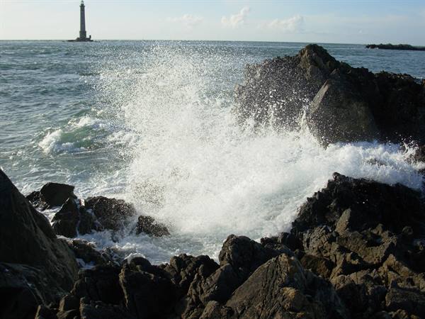 Wave crashing on black rocks, lighthouse standing in the sea behind