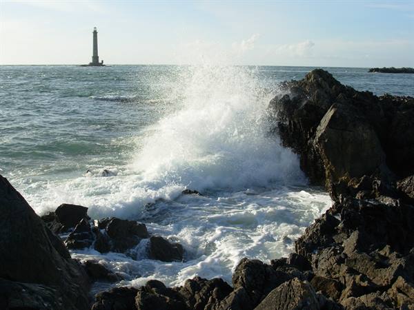 Wave crashing on black rocks, lighthouse standing in the sea behind
