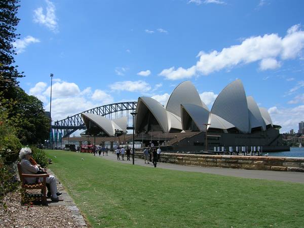 Tourists walk by the Sydney Opera House, Harbour Bridge behind, elderly woman on bench