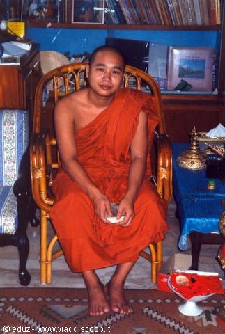 Young Buddhist monk in orange robe sitting on a rattan armchair indoors