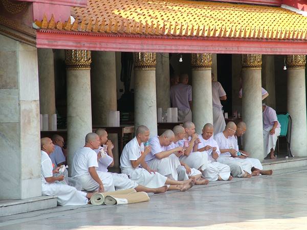 Monks in white sit eating beneath marble columns of a golden-roofed temple