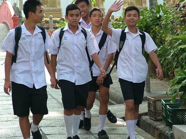 Thai schoolboys in white uniforms walk together, one waving, temple behind