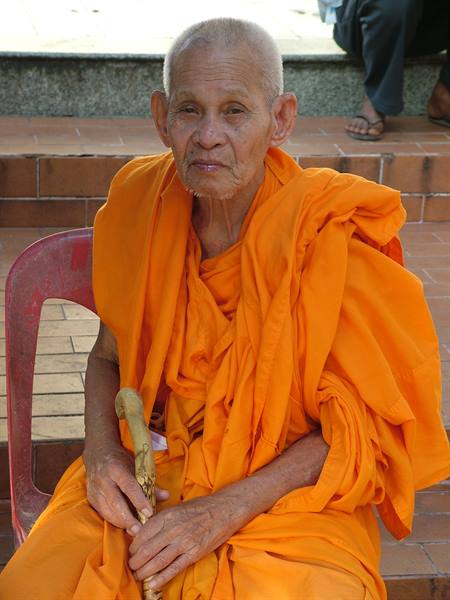 Elderly monk in orange robes sitting on a plastic chair, holding a wooden cane