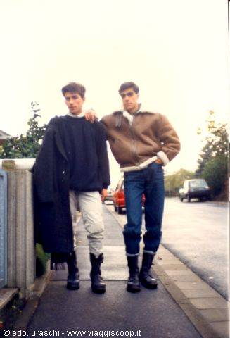 Two young friends in jackets and boots pose on a sidewalk
