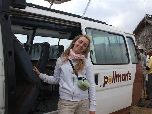 Girl beside an open Pollman's minibus, cloudy sky, thatched huts nearby