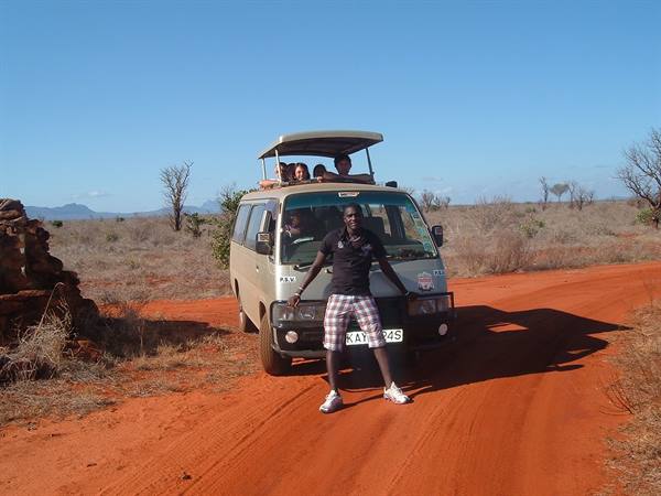Guide posing in front of safari van on red dirt road, tourists on open roof