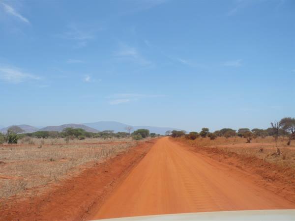 Red dirt road crossing savanna with acacias, blue mountains ahead