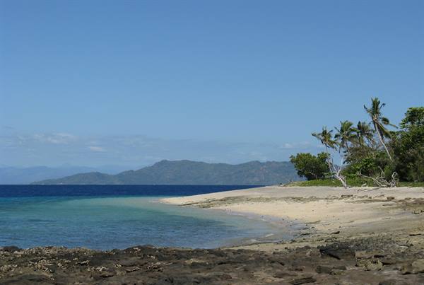 Sandy beach with palm trees, volcanic rocks in foreground and mountainous island across the channel