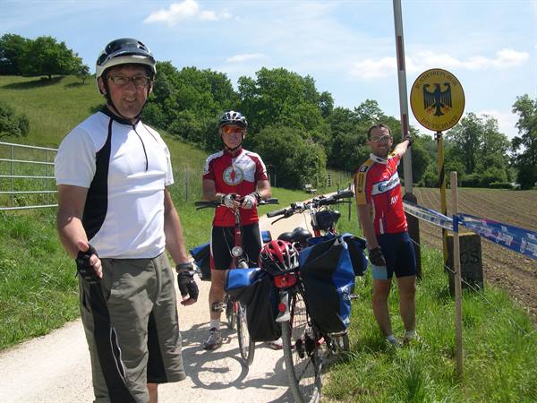 Three cyclists with loaded bikes beside a German border gate on rural road