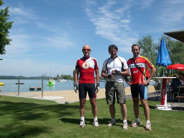 Three cyclists in cycling jerseys pose on grass by the lakeside beach