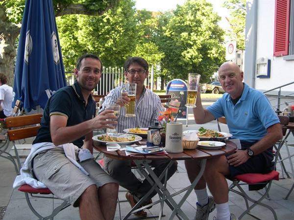 Three men toast with beers at an outdoor beer garden table