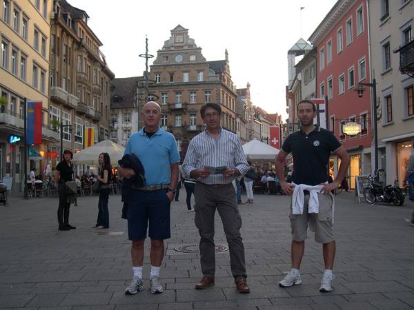 Three men pose on a cobbled square with colorful buildings and white tents