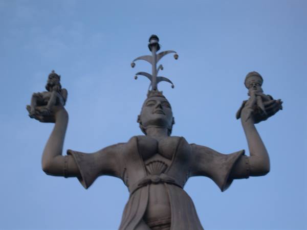 Stone statue of a woman with raised arms holding figures against blue sky