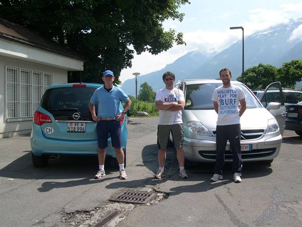 Three men beside two parked cars, snowy mountains and cloudy sky