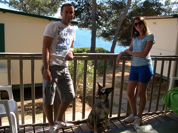 Couple with dog on wooden terrace in front of bungalows, sea behind pine trees