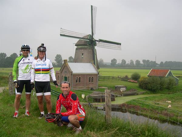 Three cyclists in cycling jerseys stop in front of a windmill