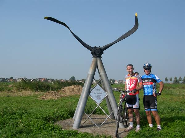 Two cyclists with mountain bike beside an airplane propeller monument in a meadow