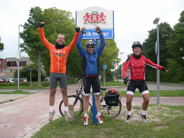 Three cyclists jump in front of the Alkmaar road sign, bike leaning on pole