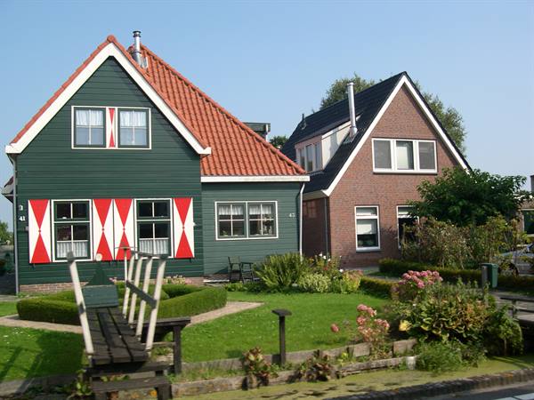 Green wooden house with red shutters and small bridge, next to brick house