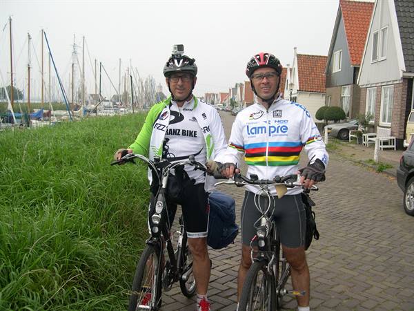 Two cyclists in helmets and racing jerseys stop their bicycles on a cobbled path beside tall grass