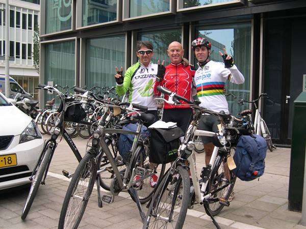 Three cyclists in racing jerseys wave among bicycles in front of a glass building