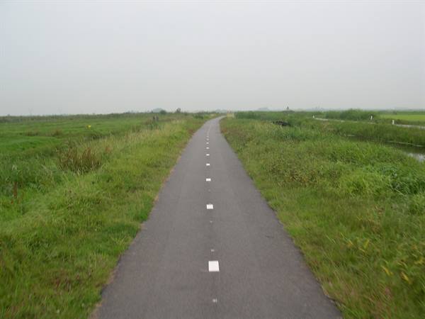Paved bike path with white markings between grassy meadows and water ditches, gray sky