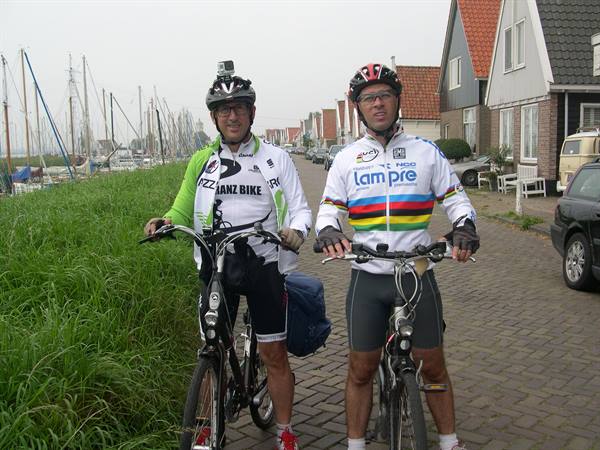 Two cyclists in racing jerseys pose with bikes on cobblestone road near marina