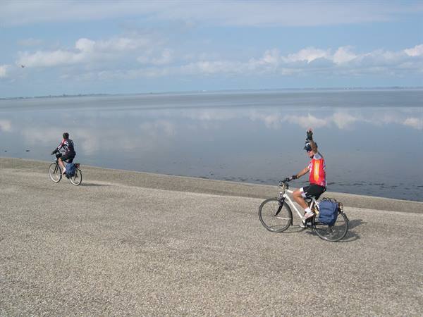 Two cyclists ride along concrete dike beside calm sea and cloudy sky