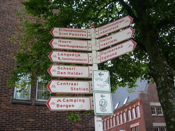 Red bicycle signpost on white pole beside brick buildings and green trees