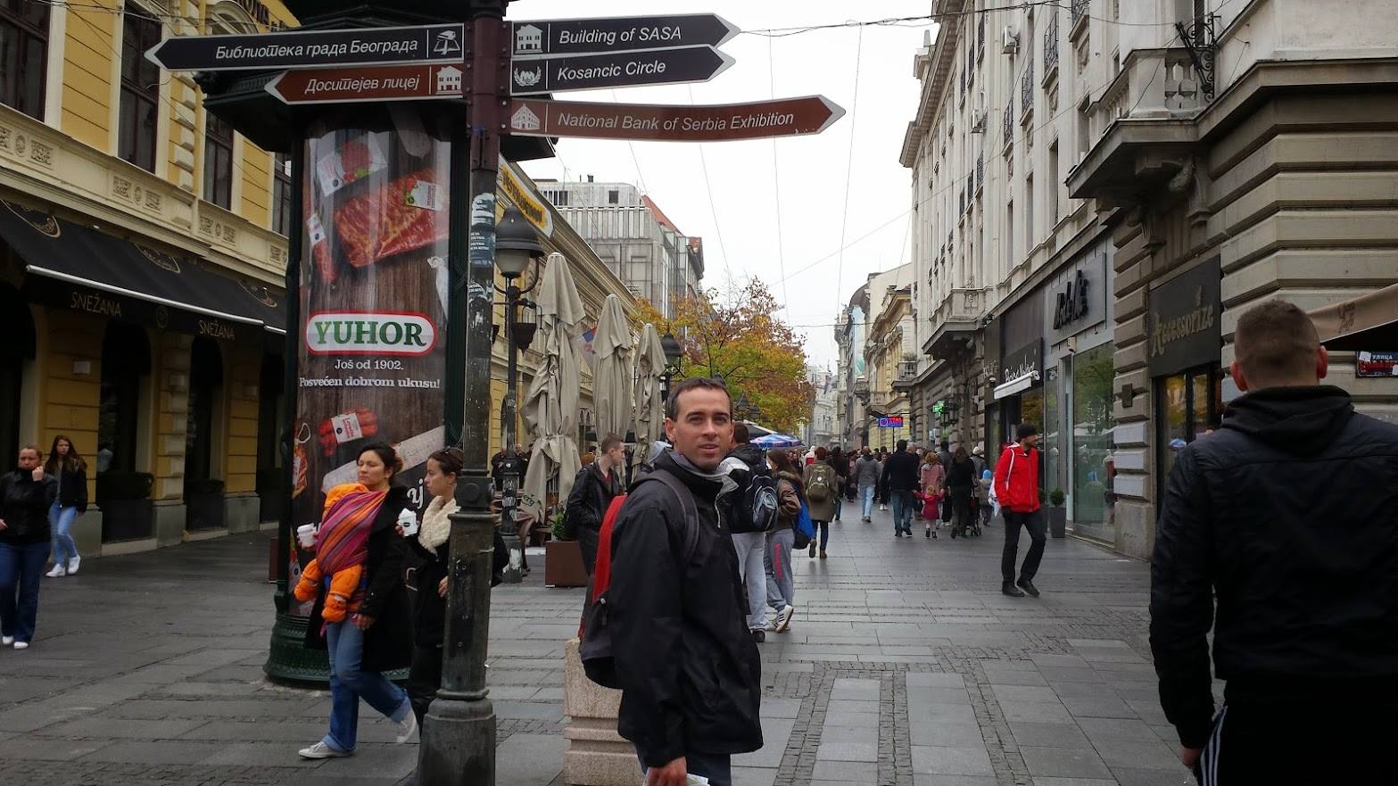Uomo con zaino su strada pedonale affollata, insegne e cartelli direzionali a Belgrado