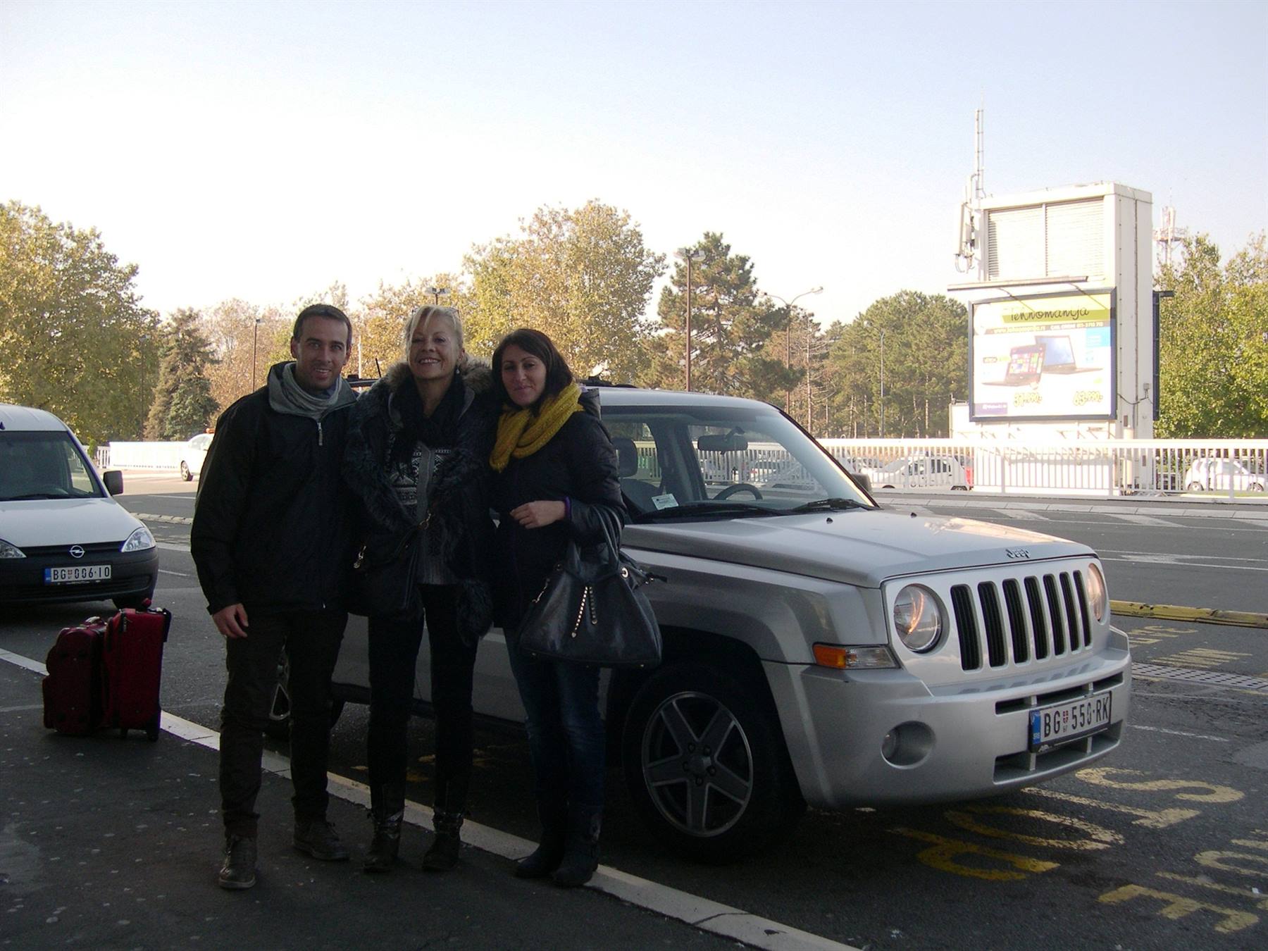 Tre amici accanto a un Jeep grigio con valigie rosse su un viale alberato