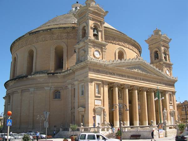 The Mosta Rotunda in golden limestone, with bell towers and colonnade under blue sky