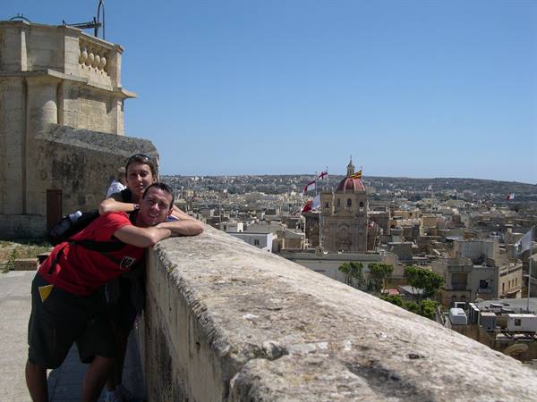 Two tourists leaning on stone ramparts in Victoria, Gozo, with church and flags