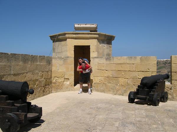 Man carrying woman among old cannons at Gozo Citadel, limestone walls, bright sun