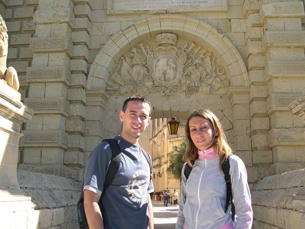 Two backpackers posing under the carved limestone gateway of Mdina