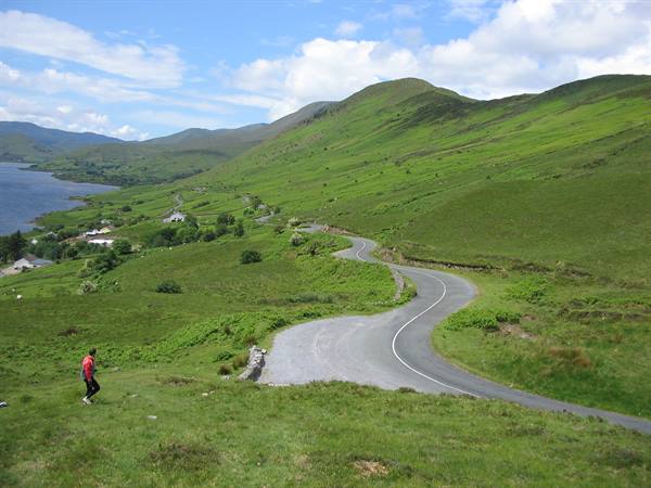 Winding road through green hills, hiker in pink walking down the grass